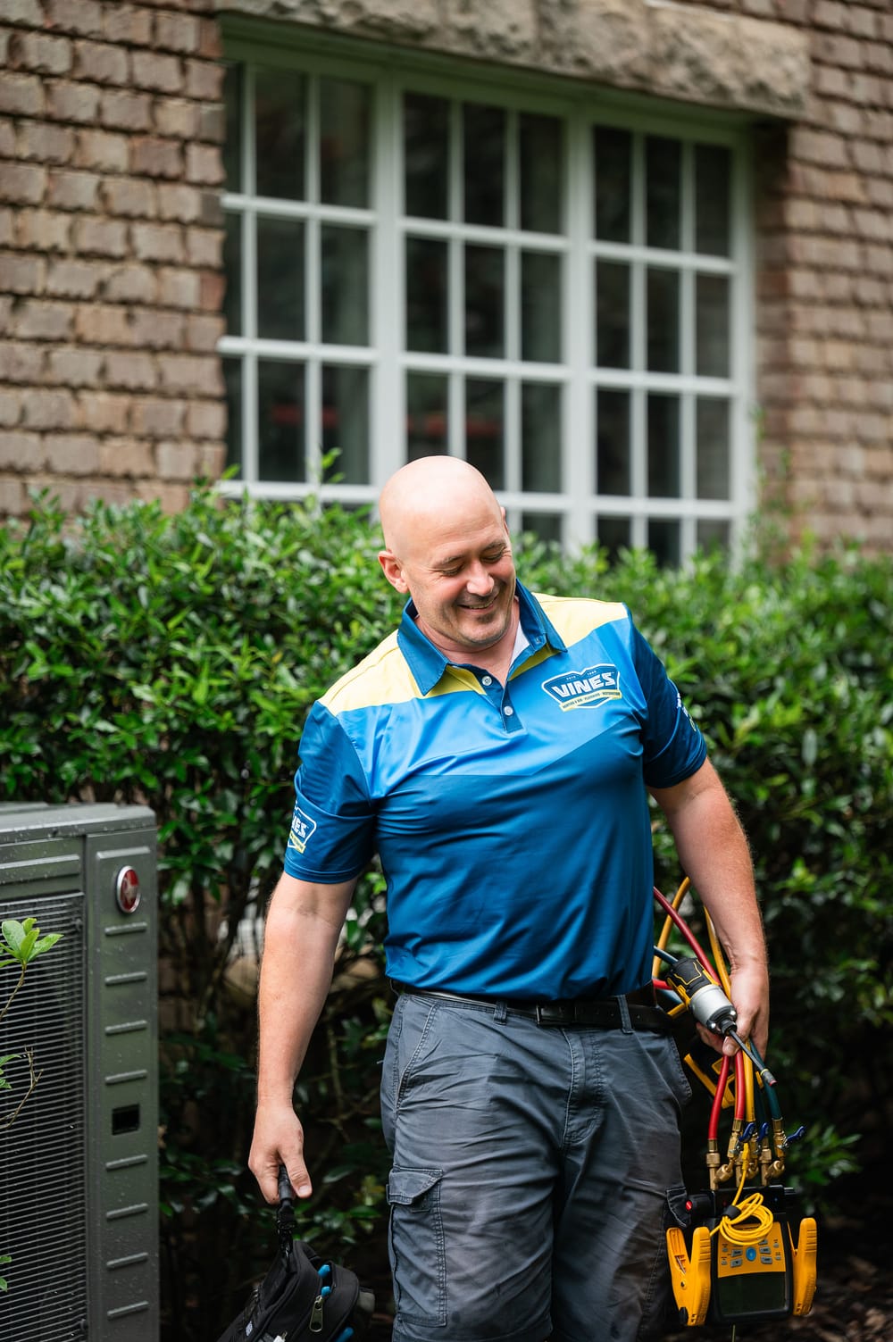 HVAC technician walking away from an outdoor air conditioning unit while carrying diagnostic tools at a residential property