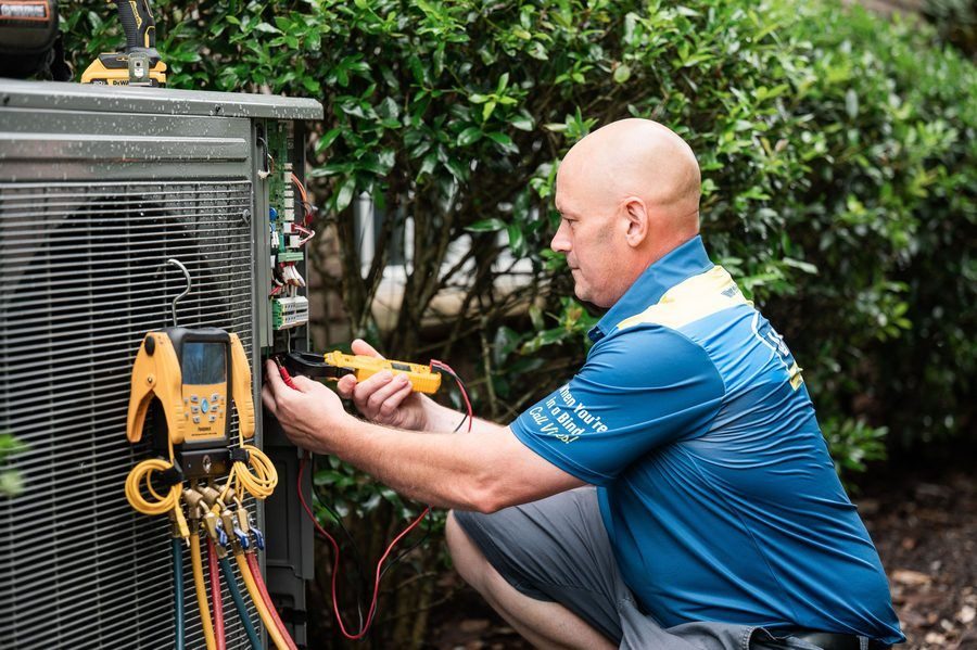 Technician in blue shirt using a multimeter to diagnose an outdoor AC unit outdoors, with gauges and cables connected to the panel.
