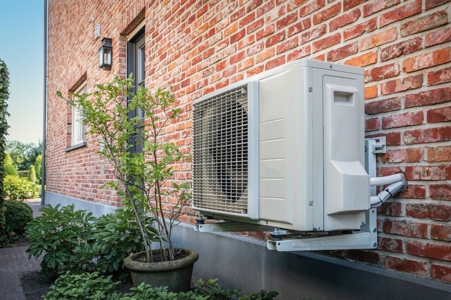 Heat pump on the side of a brick house surrounded by greenery