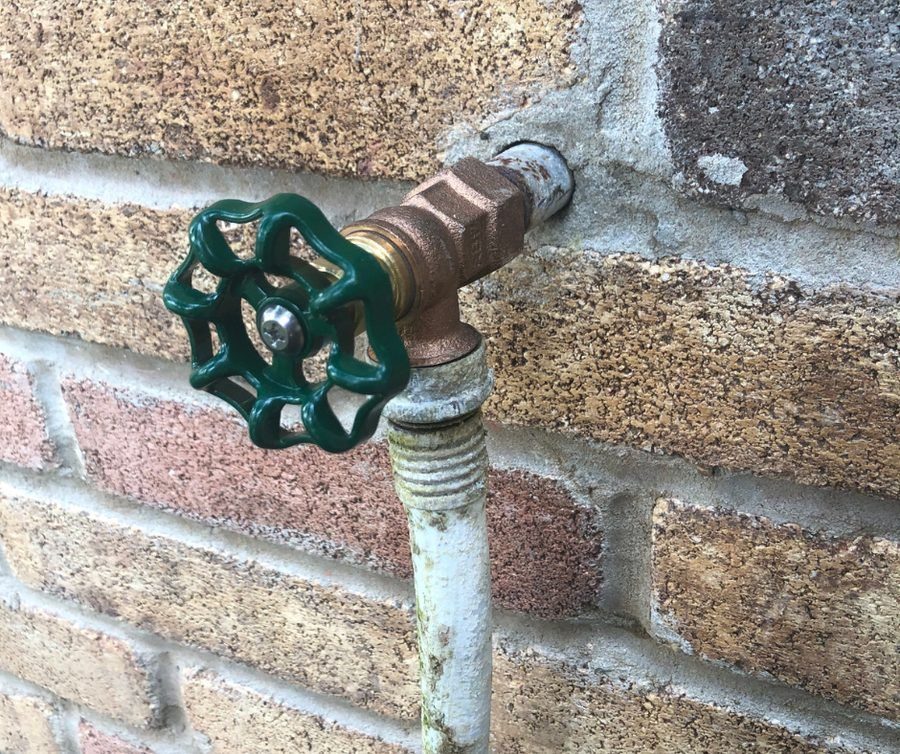 Outdoor water spigot with a green wheel handle mounted on a brick wall; shows rust and weathering.